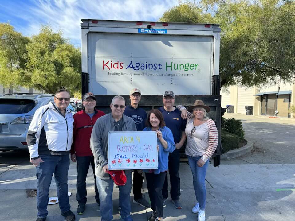 A group of eight people stands in front of a truck with "Kids Against Hunger" written on it. They hold a poster that reads "AREA 4 ROTARY-GKI 15332+ Meals!" They are outdoors on a bright day with trees and buildings in the background.