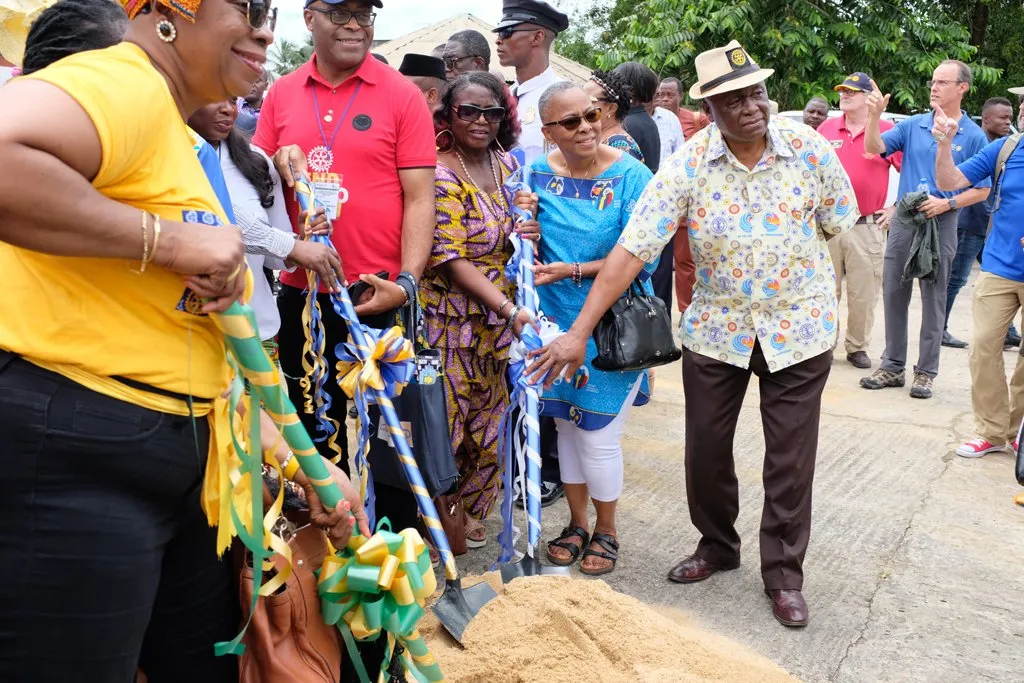 A diverse group of people gather outdoors for a ceremonial groundbreaking event. Several individuals hold decorated shovels tied with green, yellow, and blue ribbons, while others look on, smiling. Trees and other attendees are visible in the background.