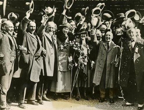 A group of men and women in vintage clothing cheerfully raise their hats and smile for the camera. Some women are holding flowers. The image has an old-time feel, suggesting it was taken in the early 20th century during a celebratory event.