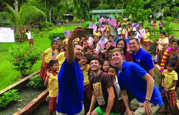 A group of adults and children are gathered outside in a lush green area, smiling and posing for a selfie. The children are holding American flags and wearing colorful clothing. In the background, there are trees, plants, and a building with a green roof.