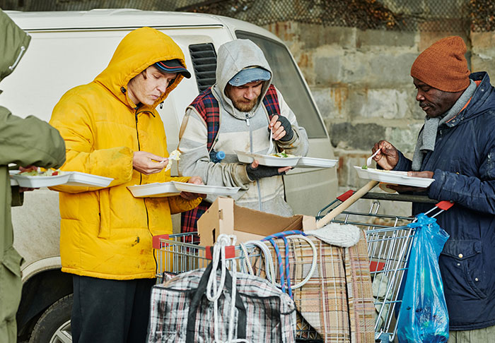 Three men in winter clothing stand outdoors near a white van, eating food from styrofoam containers. They have a shopping cart filled with bags and blankets beside them. The background features a stone wall.