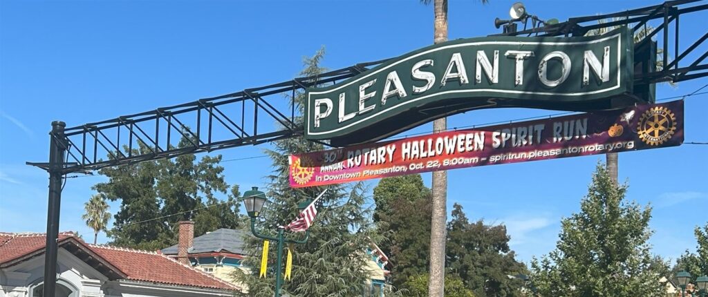 A street sign with "Pleasanton" in green letters is mounted on a supporting structure. Below it, a banner announces the "Rotary Halloween Spirit Run" event in Downtown Pleasanton on October 28 at 8:00 AM. Trees and rooftops are visible in the background.