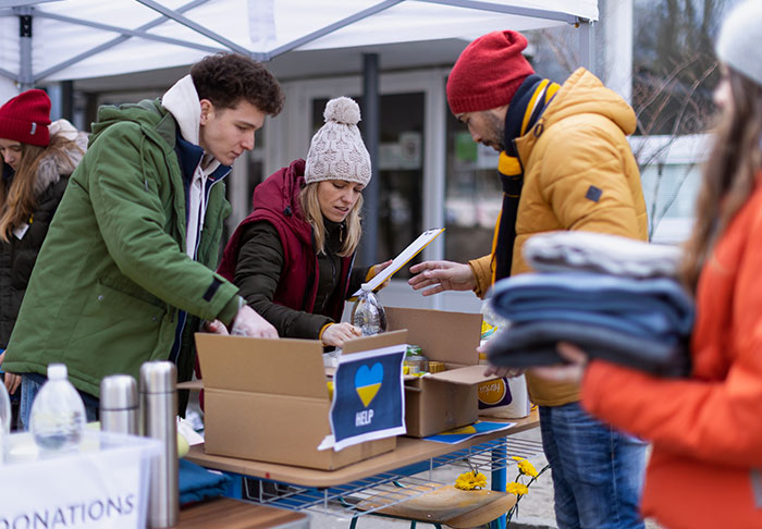 A group of people at an outdoor donation station sorting through boxes under a tent. One person is holding a pile of clothes, while others are organizing various items. A sign with a heart and the word "HELP" is visible on one of the boxes.